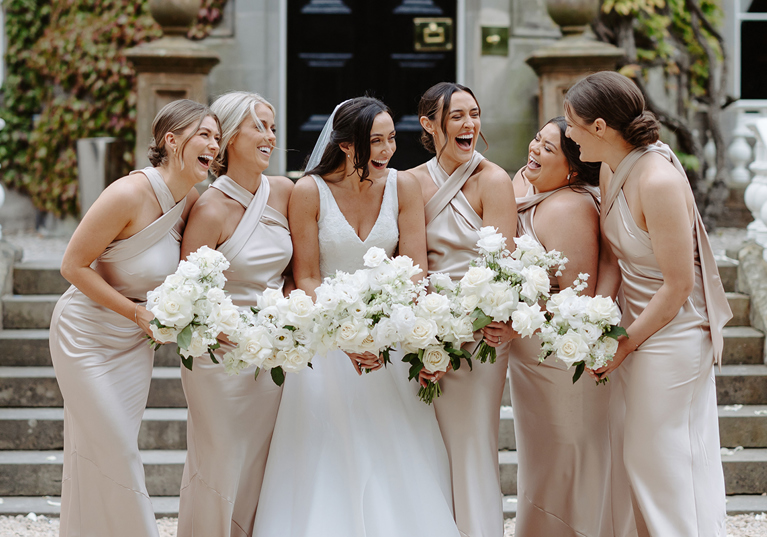 Bride and her five bridesmaids laugh together while holding white rose bouquets